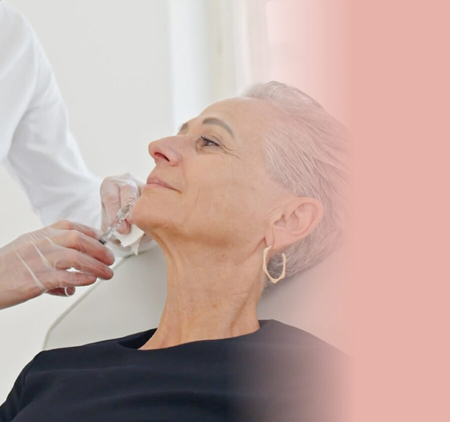 Eine Hand mit medizinischem Handschuh und Spritze, die einer lächelnden älteren Frau eine Spritze am Kinn setzt.